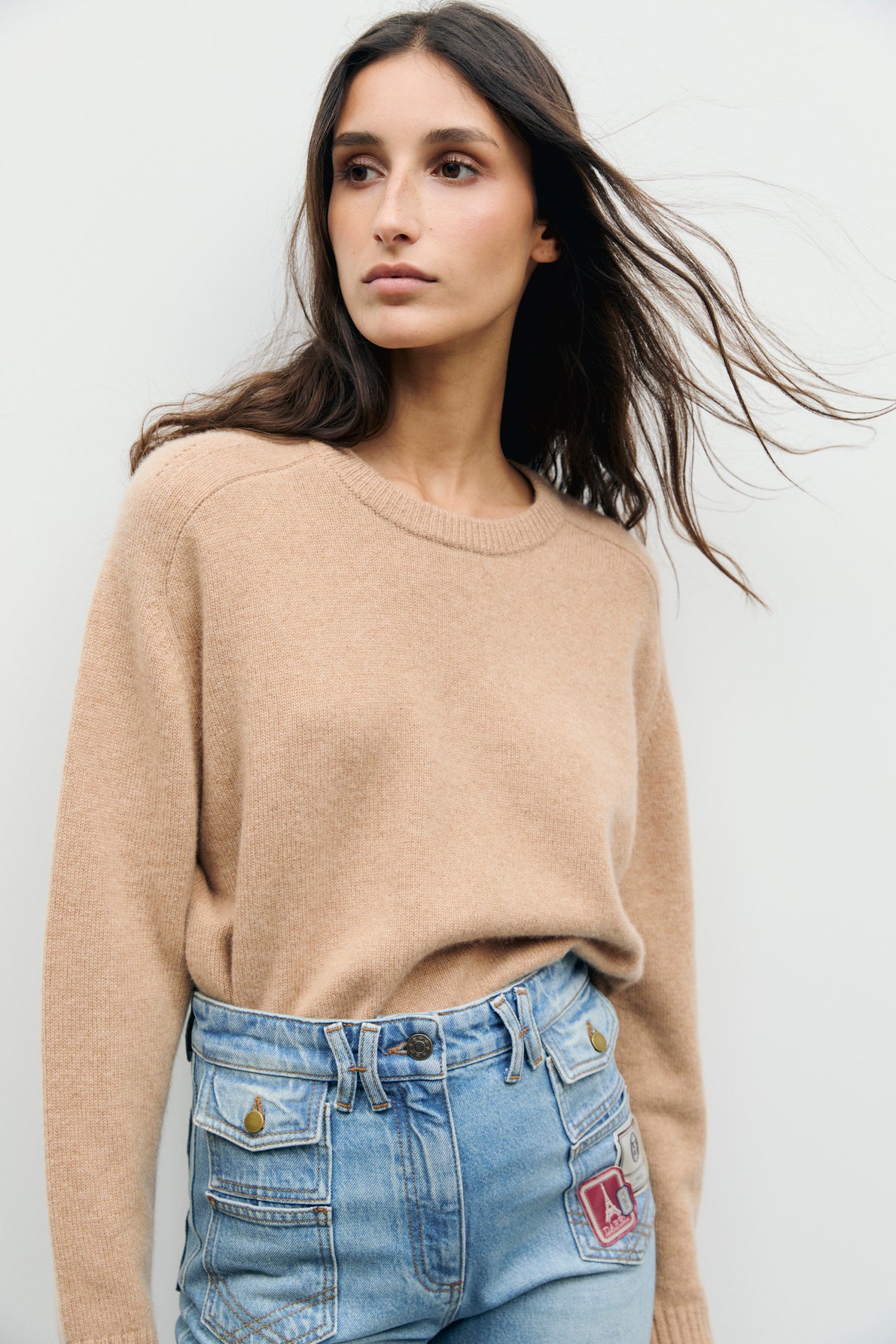 A woman with long brown hair wears a tan pull col rond in cachemire, tucked into high-waisted blue jeans with patches. She stands against a plain light background, looking to the side with a calm expression.