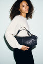 A woman with curly hair, wearing a pull écru sweater and black pants, holds a large black leather handbag against a plain light background.