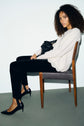 A woman with curly hair sits sideways on a wooden chair, wearing a pull laine femme in a soft white shade, along with black pants and black heels. She holds a black bag on her lap and looks toward the camera with a neutral expression.