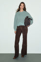 A woman stands against a plain background wearing a light green pull laine femme with a scalloped hem, brown suede pants, brown boots, and holding a dark brown bag.