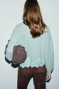 A person with long brown hair, wearing a light blue pull dentelle sweater with lace trim and brown pants, holds a brown drawstring bag. The image is taken from behind.