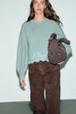 A woman wearing a pull laine femme in light blue with delicate lace trim and brown pants holds a textured brown handbag while walking against a light background.