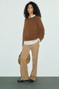 A woman with curly hair wears a brown pull col rond sweater over a white shirt, tan pants, and black shoes, holding a tan bag, standing against a light background.