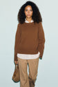 A woman with curly hair wears a brown pull laine sweater over a white shirt and tan pants, holding a brown suede handbag, standing against a light blue background.
