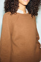 A woman with curly hair wears a light brown pull laine knit sweater over a striped shirt. The image focuses on her upper torso and part of her face, shown against a pale background.
