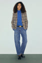 A person with curly hair wears a plaid blazer, a pull col roulé bleu in laine mérinos, loose blue jeans, black belt with a round buckle, and black shoes, standing with hands in pockets against a plain background.