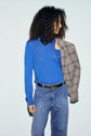 Woman with curly hair wearing a pull bleu col roulé en laine mérinos and jeans, holding a plaid blazer over her shoulder, looking to the side against a light background.