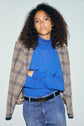 A person with curly hair wears a pull col roulé bleu en laine mérinos, a plaid blazer draped over their shoulders, and blue jeans, posing against a light background.