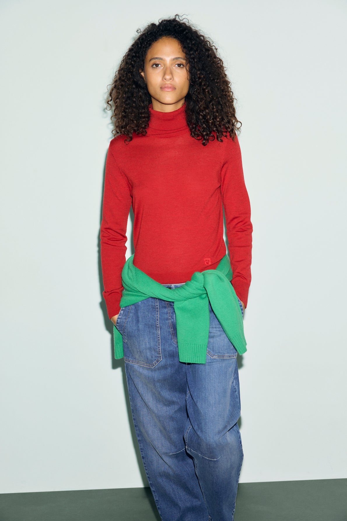 Woman with curly hair wears a pull col roulé rouge vibrant, oversized blue jeans, and a green sweater tied around her waist, standing against a plain light background.