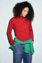 A person with curly hair wears a pull col roulé rouge vibrant and blue jeans, with a green sweater tied around their waist. Made from douce laine mérinos, the sweater stands out as they look down with hands in pockets, standing against a light background.