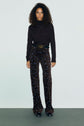 A person with curly hair wears a pull col roulé en laine mérinos, brown high-waisted pants with small blue prints, a wide belt, and black shoes, standing against a plain light background.