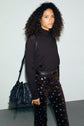 Woman with curly hair in a pull col roulé noir, brown pants with blue flower print, and holding a black fringed bag, standing against a plain light background.