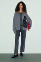 A person with curly hair wears a gilet gris with a brown col fausse fourrure, gray jeans, and black loafers, holding a black purse and colorful scarves against a plain light background.