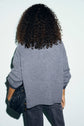 A person with curly hair, seen from behind, is wearing a loose gilet gris and dark jeans. They are holding a textured black handbag in their left hand. The cozy ensemble suggests a hint of cachemire warmth, set against a plain and light-colored background.