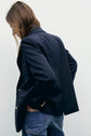 A person with long brown hair wears a blazer bleu marine croisé doré and blue jeans, standing sideways against a plain light background, looking down.