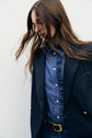 A person with long brown hair wearing a blue ruffled shirt, a blazer bleu marine, and a black belt with a gold buckle stands against a plain light background, looking downward.