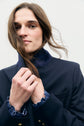 Person with long brown hair in a blazer bleu marine and shirt holds a strand of hair across their face, looking at the camera with a neutral expression against a plain light background.