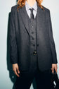 A person wearing a dark pinstripe suit with a matching veste rayée, striped shirt, and dark tie is holding a bag in one hand. The person’s head is partly out of the frame. The background is a plain light color.