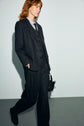 Person with shoulder-length hair wears a dark veste rayée suit, vest, and tie, walking against a plain light background while holding a small handbag.