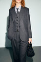 A person wearing an anthracite pinstripe suit, complemented by a matching vest and tie, holds a black handbag. The background is plain, and the person’s face is partially out of the frame.