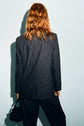 Person with wavy, shoulder-length reddish hair stands facing away, wearing a dark veste rayée and holding a small black handbag against a light background.