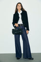 Woman standing against a plain wall, wearing a veste courte noire with boutons dorés, a white blouse with a bow, dark wide-leg jeans, black shoes, and holding a black handbag.