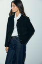Woman wearing a veste courte noire in tweed bouclé with boutons dorés over a white blouse with a necktie and wide-leg dark jeans, standing against a plain light wall, looking to the side with one hand in her pocket.