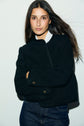 A woman with long dark hair wearing a veste courte noire in textured tweed bouclé, adorned with boutons dorés, stands against a plain light background, looking directly at the camera with a neutral expression and her arms crossed.