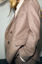 A person with long blond hair wears a light brown veste de tailleur and a striped shirt, with one hand in their pocket. The image is cropped to show the torso and part of the face, slightly turned to the side.