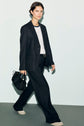 A person walks in a dark pinstripe suit with wide-leg pants, white T-shirt, and casual sneakers, carrying a black bag. This veste rayée femme look exudes effortless sophistication against the light and plain background.