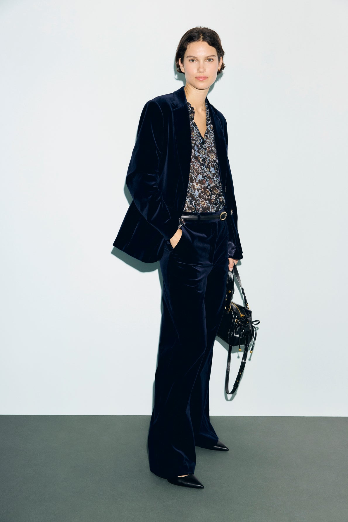 A person stands against a plain background wearing a veste tailleur velours—a dark velvet suit—paired with a patterned blouse and pointed black shoes, holding a black handbag by their side with one hand in their pocket.