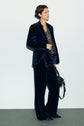 A person wearing a veste tailleur velours dark velvet pantsuit, a patterned blouse, and black pointed shoes stands against a plain light background, holding a black handbag.