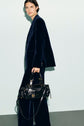 A person in a navy blue veste tailleur velours stands holding a black leather handbag with gold hardware. The background is a plain light gray wall and gray floor.