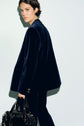 Person with short dark hair wearing a navy velvet suit tailleur velours looks over their shoulder, holding a large black handbag, standing against a plain light background.