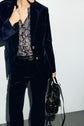 A person wearing a dark blue veste tailleur velours suit, a floral blouse, and a black belt holds a black handbag. The face is partially out of frame against a plain, light-colored background.