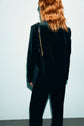 A person with wavy, shoulder-length red hair stands facing away, wearing a dark veste de tailleur en velours ras and holding a black purse with a gold chain strap. The background is light and plain.