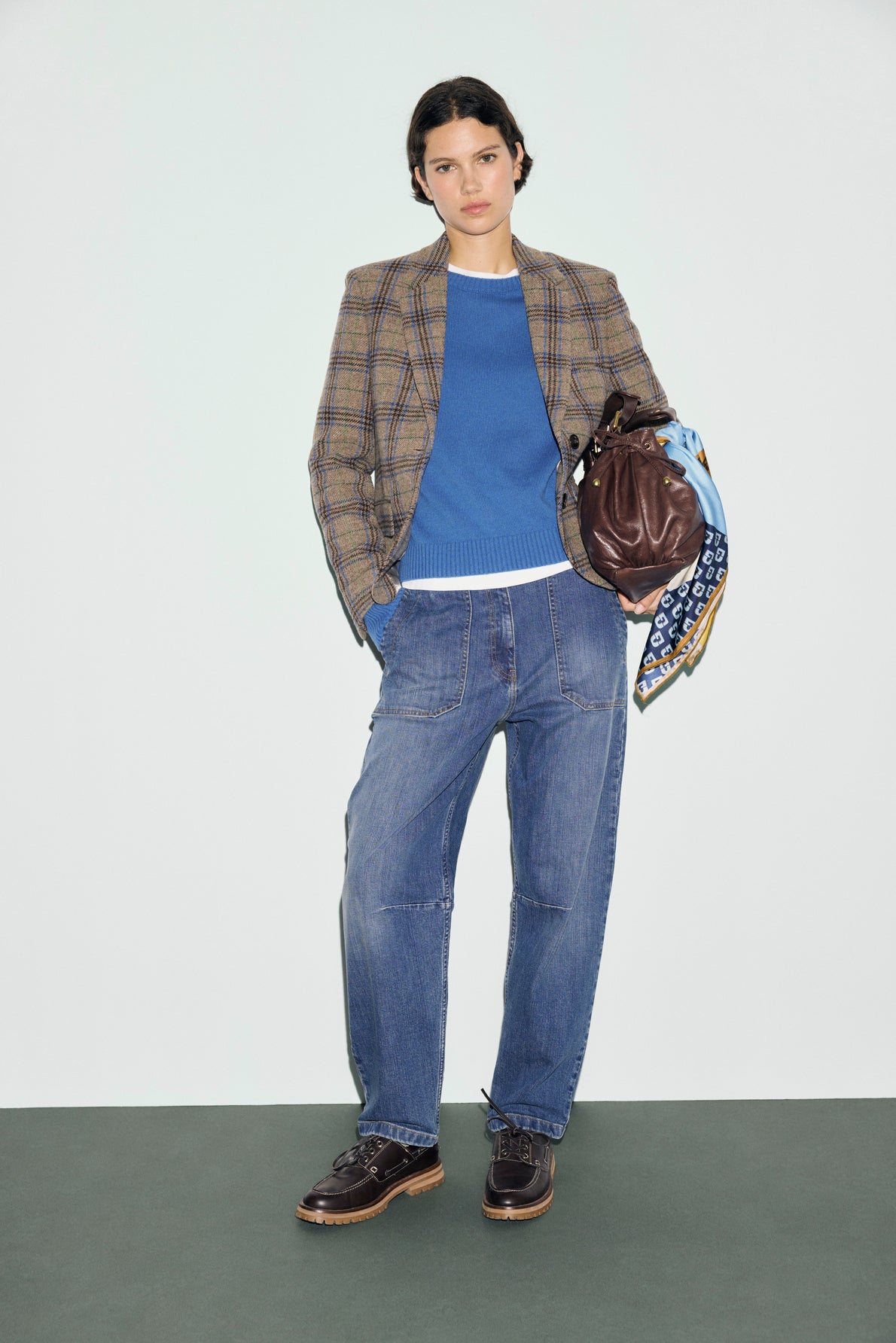 A person in a veste à carreaux, blue sweater, and loose jeans stands holding a brown leather bag with a patterned scarf tied to it. They wear dark shoes and have short, dark hair. The background is light and plain.