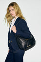A person with long blonde hair wearing a navy veste tailleur femme and jeans holds a black leather shoulder bag, posing against a plain white background.