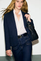 A person with long hair wears a navy veste tailleur femme blazer crafted from laine vierge, paired with a white ruffled blouse, blue jeans, and a black belt, holding a black leather shoulder bag, standing against a plain white background.