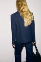 Person with long blonde hair stands facing a wall, wearing a dark blue veste tailleur femme and matching pants, holding a black bag at their side. The background is plain and white.