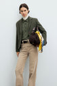 A person wearing a green veste de tailleur in laine vierge, paired with a green shirt, beige pants, and a white turtleneck stands against a light background, holding a brown bag with a yellow scarf tied to it.