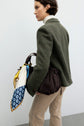 A woman in a veste de tailleur vert and beige pants carries a brown textured handbag with a colorful scarf tied to its handle. She is facing away from the camera against a plain light background.