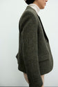 A person in a grey herringbone blazer crafted from laine vierge and a white turtleneck stands sideways against a plain light background. Only the upper body and face in profile are visible.