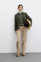 A person stands against a white wall wearing a green veste de tailleur crafted from laine vierge, paired with a light green shirt, beige pants, and brown shoes. They hold a dark bag with colorful scarves tied to it.