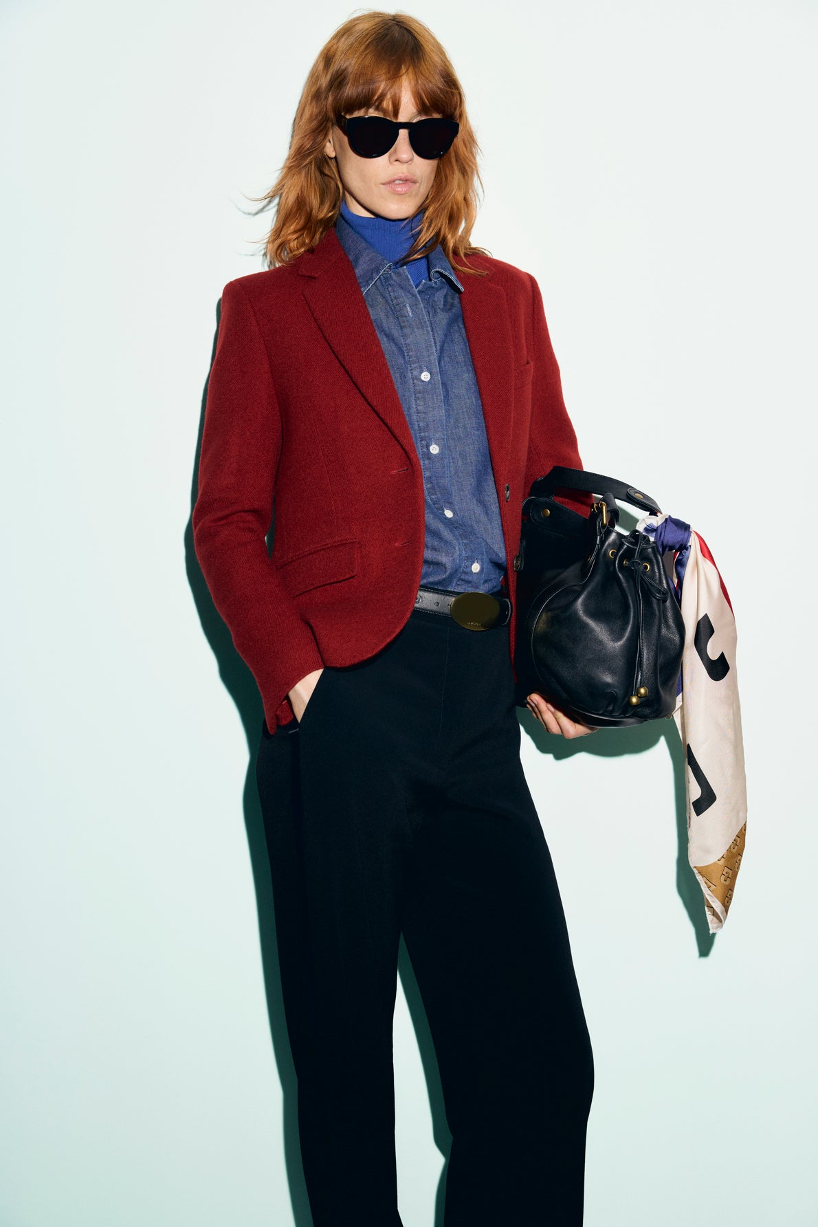 Person with red hair in sunglasses wears a veste rouge femme, styled as a vibrant red blazer, over a blue shirt and black pants, holding a black handbag with a white printed scarf against a plain light background.