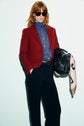 Person with red hair in sunglasses wears a veste rouge femme, styled as a vibrant red blazer, over a blue shirt and black pants, holding a black handbag with a white printed scarf against a plain light background.