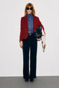 Person wearing sunglasses, a veste rouge femme—a tailored red blazer—in laine stands against a plain background, holding a black bag with a patterned scarf draped over it. The look is completed with a blue shirt and black pants for effortless sophistication.