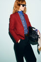 A person wearing sunglasses, a veste rouge femme styled as a red blazer, paired with a blue button-up shirt and black pants, stands against a light background, holding a black handbag with a white scarf draped over it.