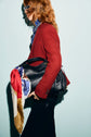 Person in a veste rouge femme and glasses holds a black handbag with a colorful scarf tied to the handle, standing against a light blue background.
