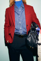 Person wearing a veste rouge femme styled as a blazer, paired with a blue shirt, blue turtleneck, and black pants, holding a black leather drawstring bag with a patterned scarf tied to it. Face is not visible.